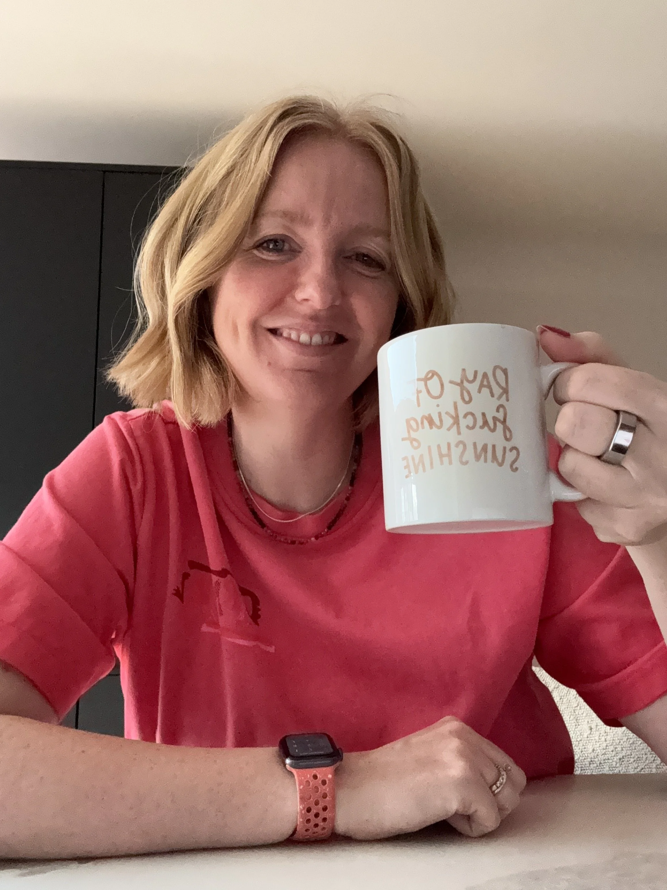 Lee smiling to camera, holding a cup which reads 'ray of fucking sunshine'