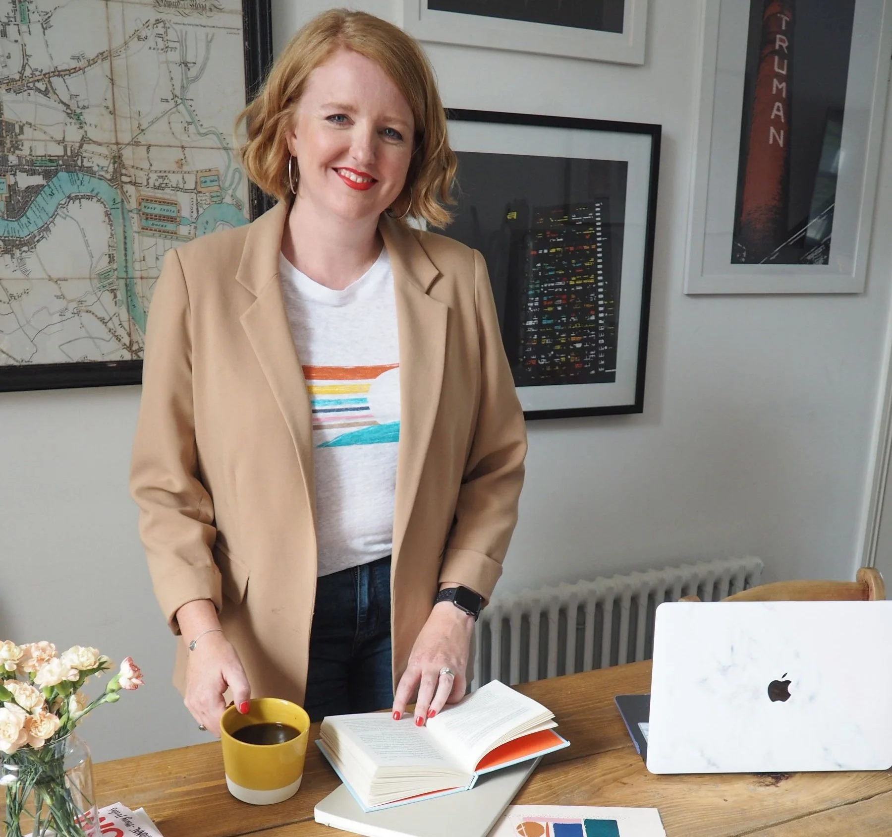 A woman with red hair, wearing a beige blazer and white T-shirt with a rainbow and sun graphic, standing at a wooden table with an open book, a yellow mug, and a marble laptop. She is smiling in a room with framed artwork and a map on the wall.