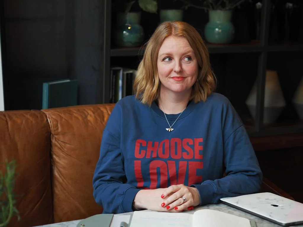 Lee Griffith sitting at a table, hands crossed in front of her on the table. She is smiling and looking up and to the right of the camera. She's wearing a choose love sweatshirt.
