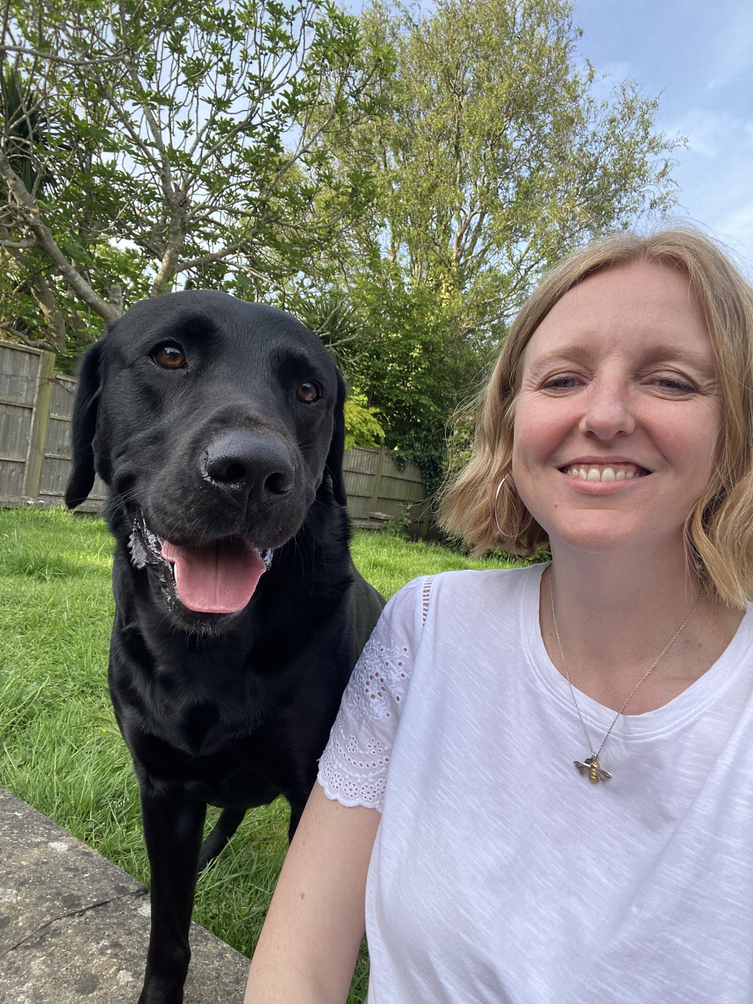 A woman smiling next to a black Labrador Retriever in a backyard with green grass, trees, and a wooden fence, under a partly cloudy sky.