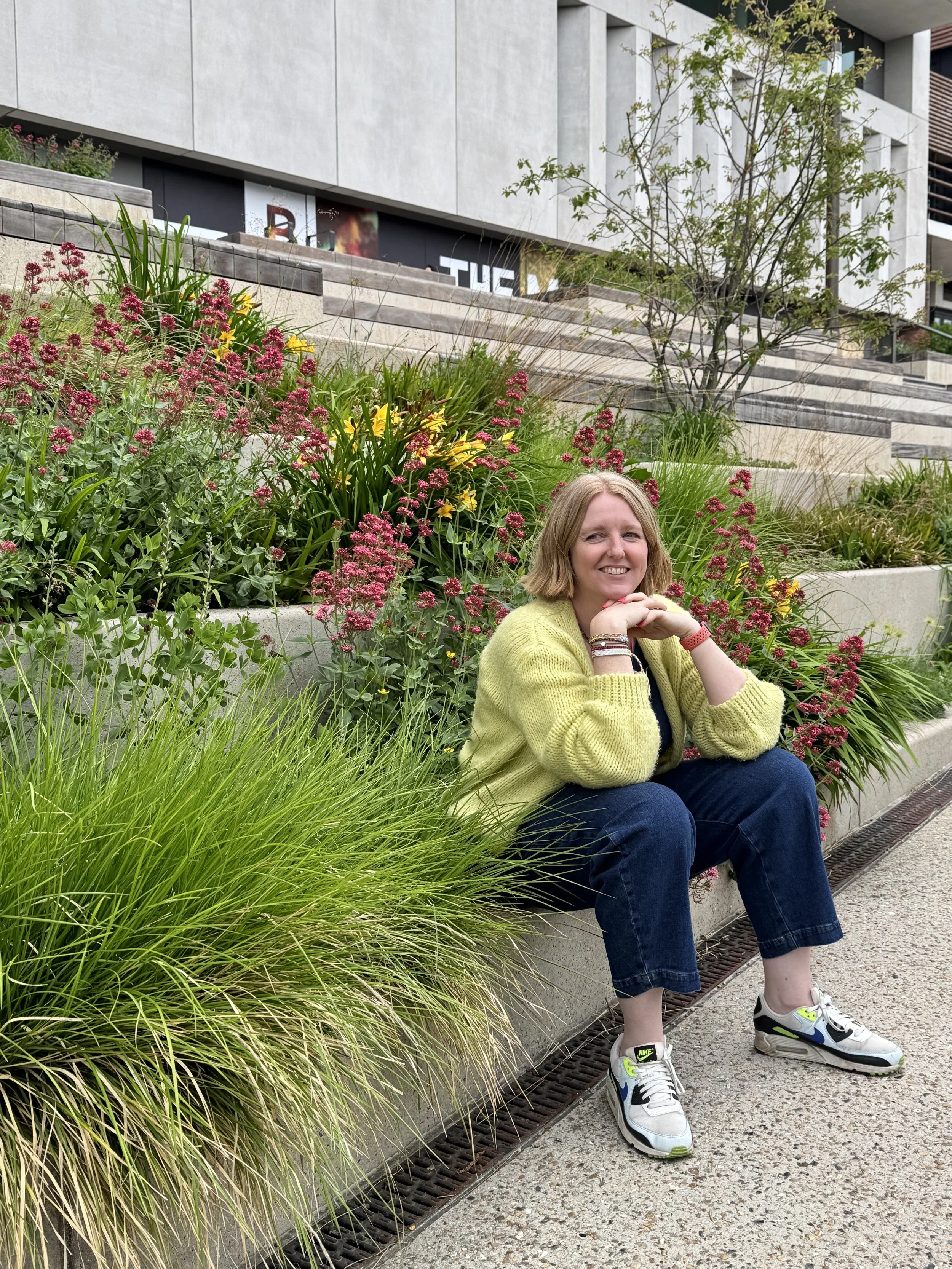 Lee Griffith, wearing denim jumpsuit and yellow cardigan, sat on concrete sleeper and is surrounded by in bloom flowers. She's looking off camera, head perched on her hands.