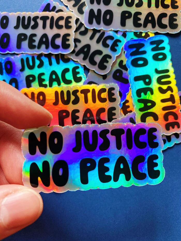 Photo of a hand holding a holographic vinyl sticker that reads "no justice no peace" set against a blue background.