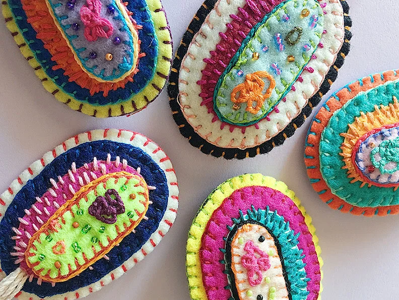 Detail group photo of 5 hand-sewn, beaded and embroidered brooches in the shape of a bacteria cell on a white background.
