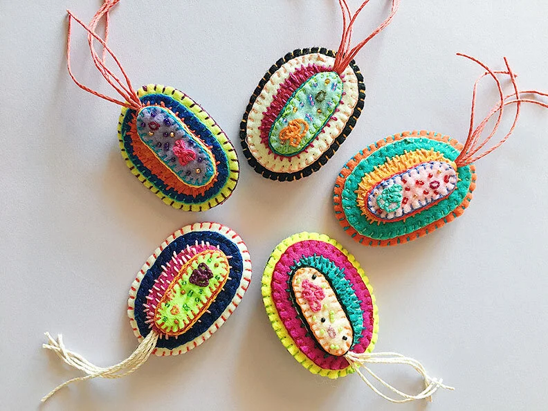 Overview group photo of 5 hand-sewn, beaded and embroidered brooches in the shape of a bacteria cell on a white background.