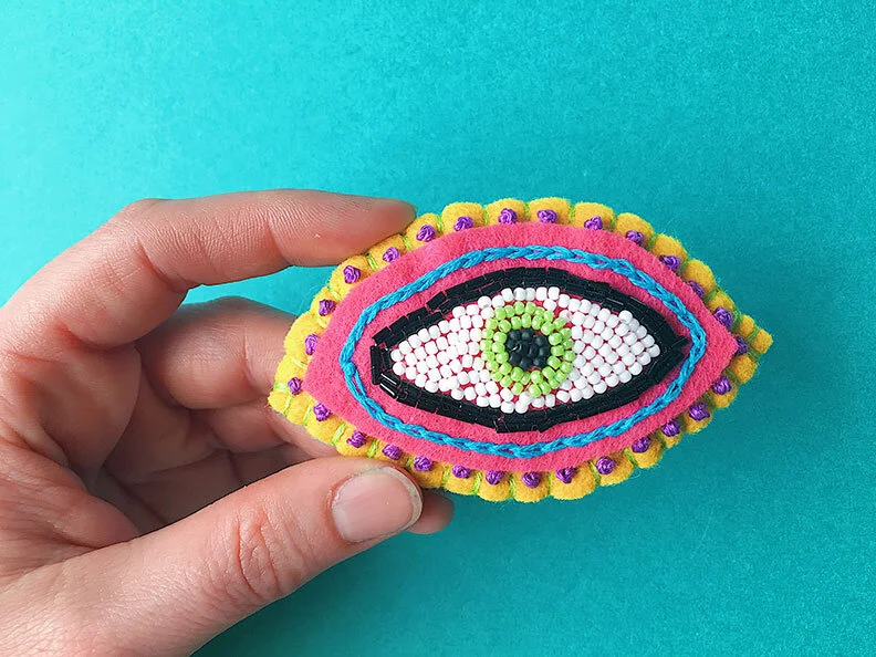 Overview photo of a hand holding a hand-sewn, beaded & embroidered brooch in the shape of an eye on a teal background.
