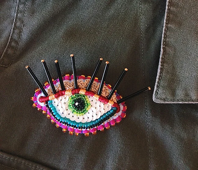 Overview photo of a hand-sewn, beaded & embroidered brooch in the shape of an eye with long bead eyelashes on a green jacket.
