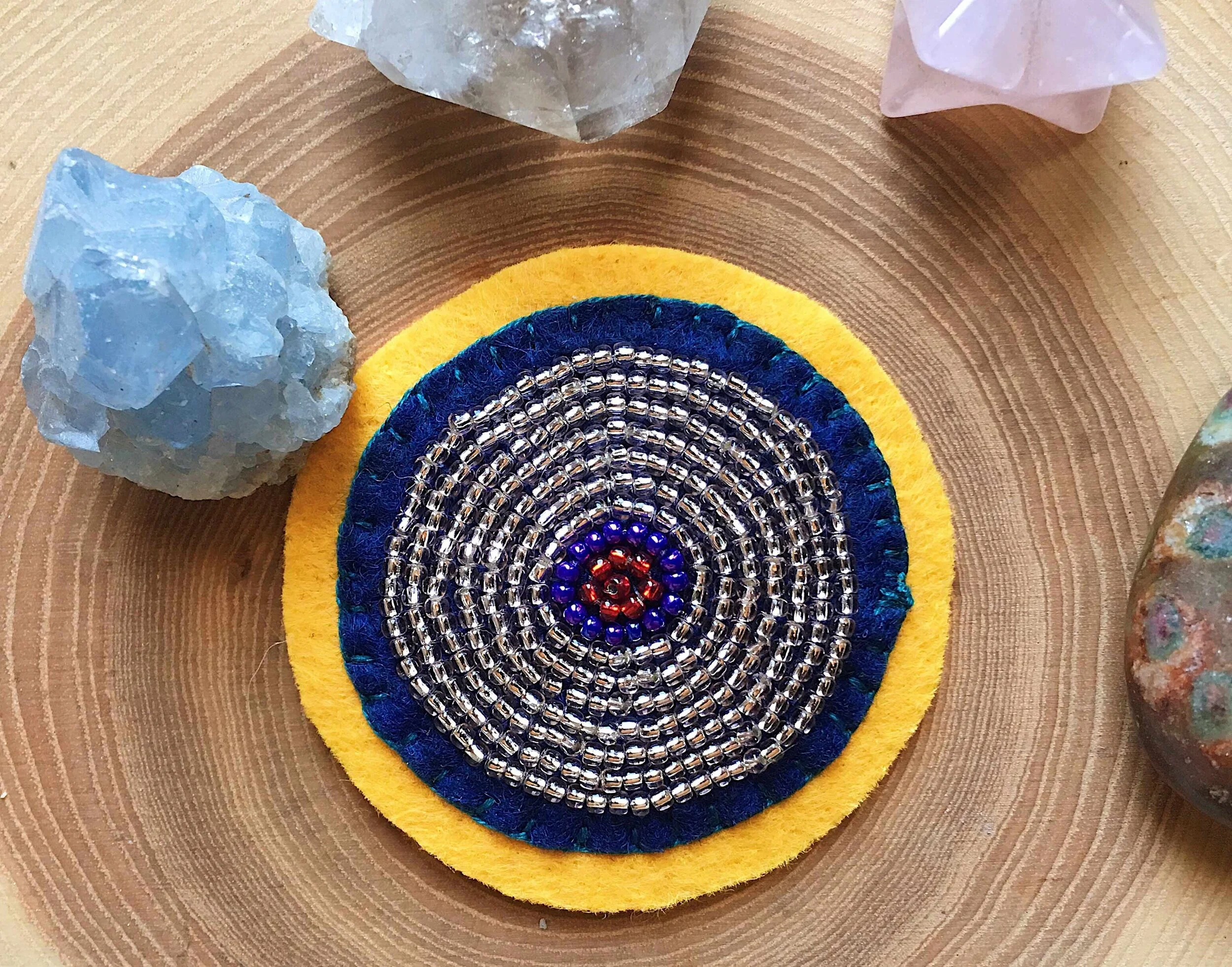 Close up photo of a hand beaded and embroidered patch of a boob, or tata, on a wooden board surrounded by crystals.