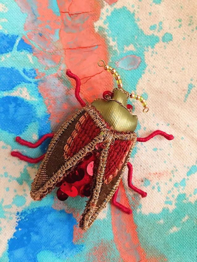 Close up photo of a hand sewn beetle art brooch with red sequins and faux gold leather on a colorful painted background.