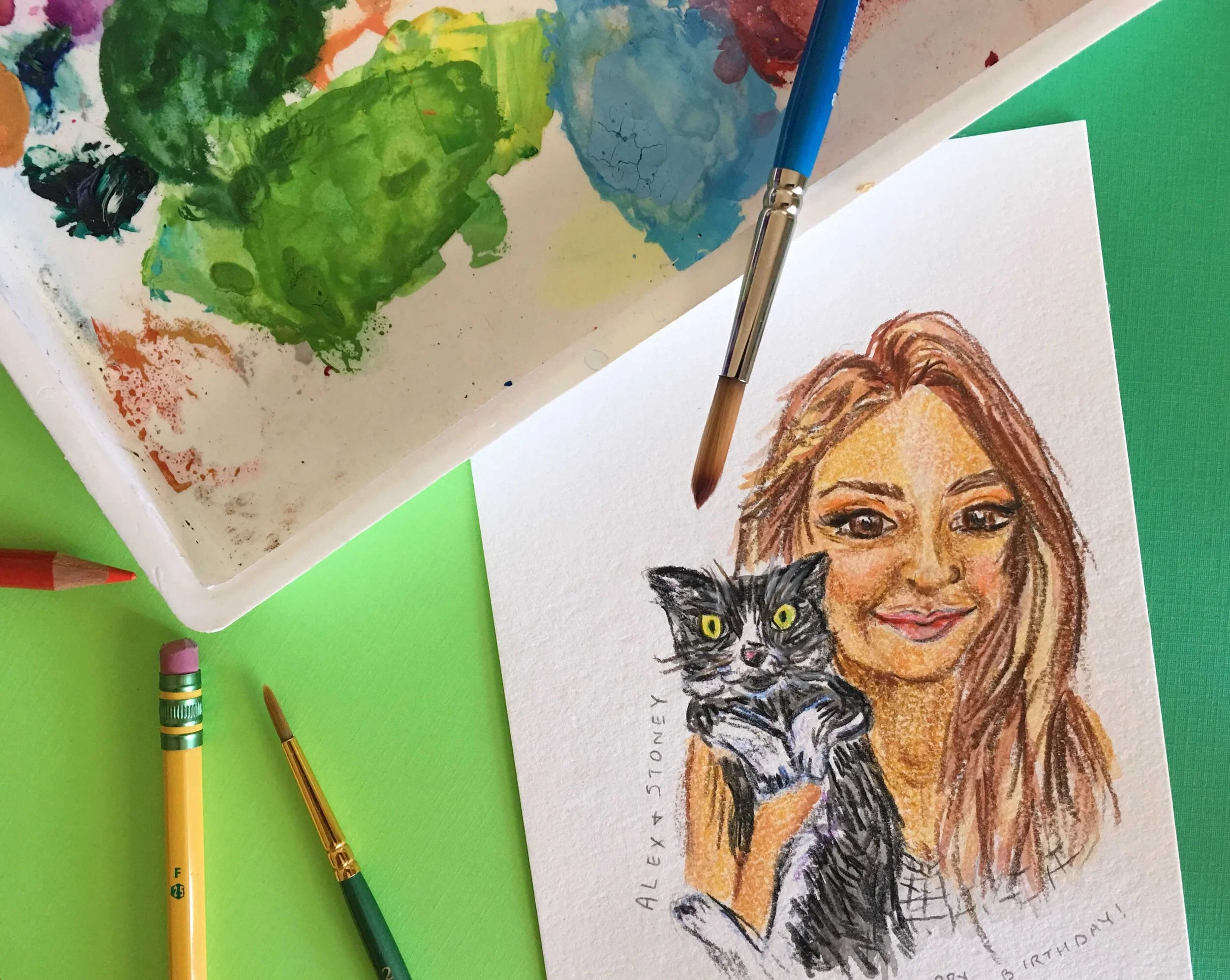 Photo of art supplies surrounding a portrait of a woman holding a cat using gouache and colored pencil.
