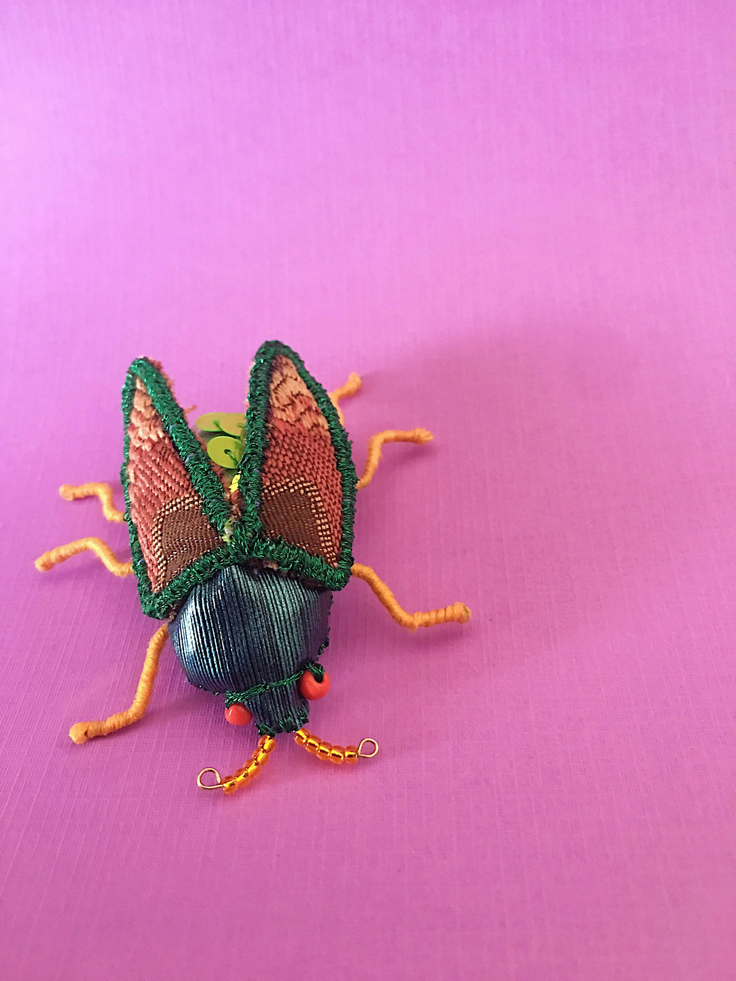 Close up photo of a hand sewn beetle art brooch with green sequins and faux blue leather on a purple background.