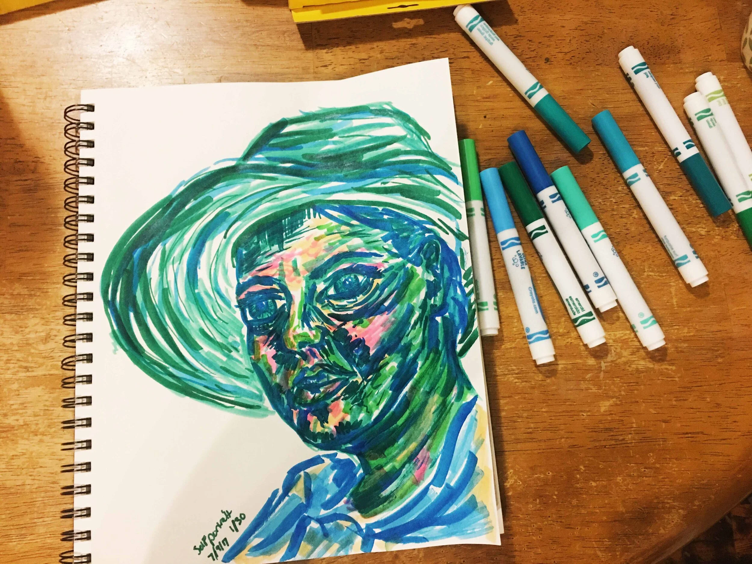 Day 1: photo of a self-portrait wearing a hat using markers surrounded by art supplies on a table.