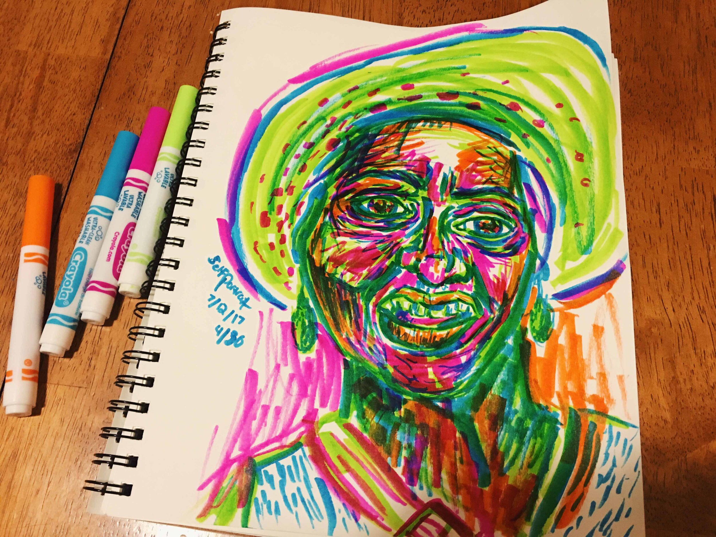 Day 4: photo of a self-portrait wearing a hat using markers surrounded by art supplies on a table.