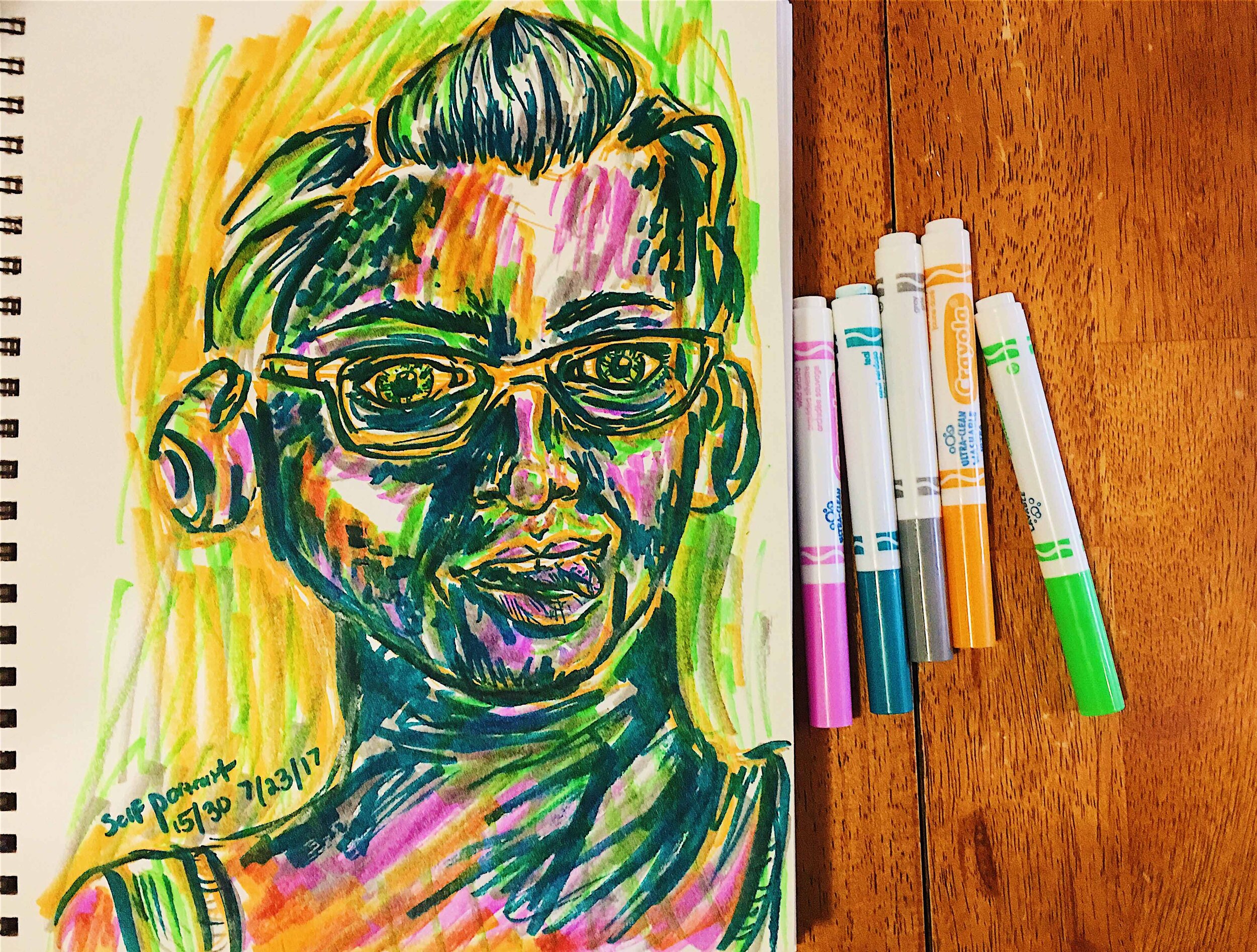Day 15: photo of a self-portrait wearing glasses and sticking my tongue out surrounded by art supplies on a table.