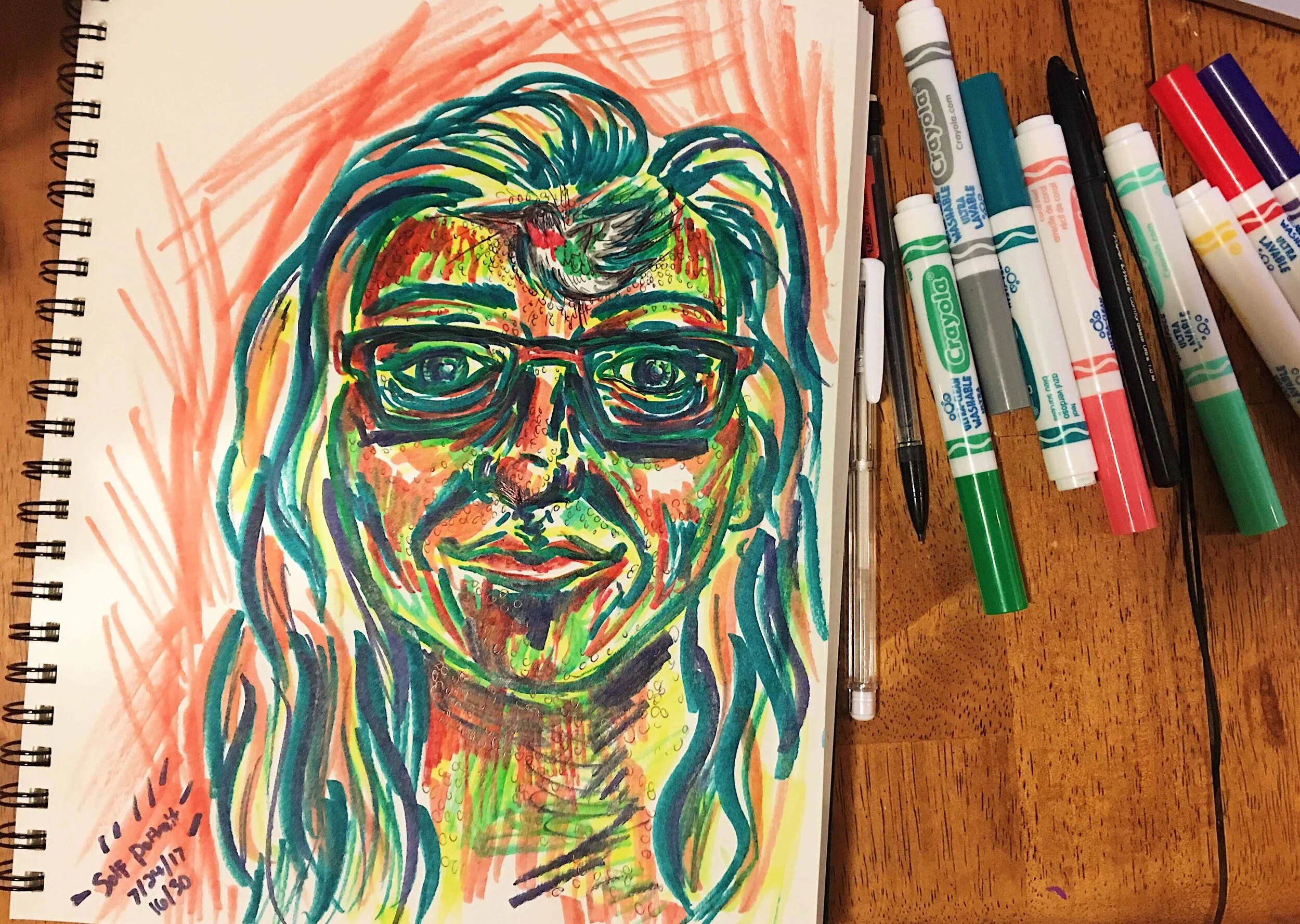 Day 16: photo of a self-portrait wearing glasses and a hummingbird surrounded by art supplies on a table.