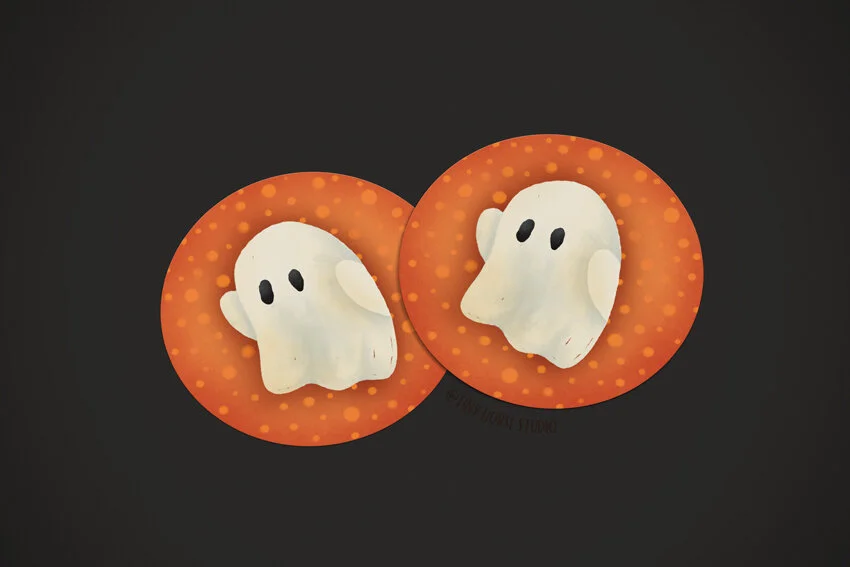 Overview photo of two round vinyl stickers that have a cute little ghost illustration set against a black background.