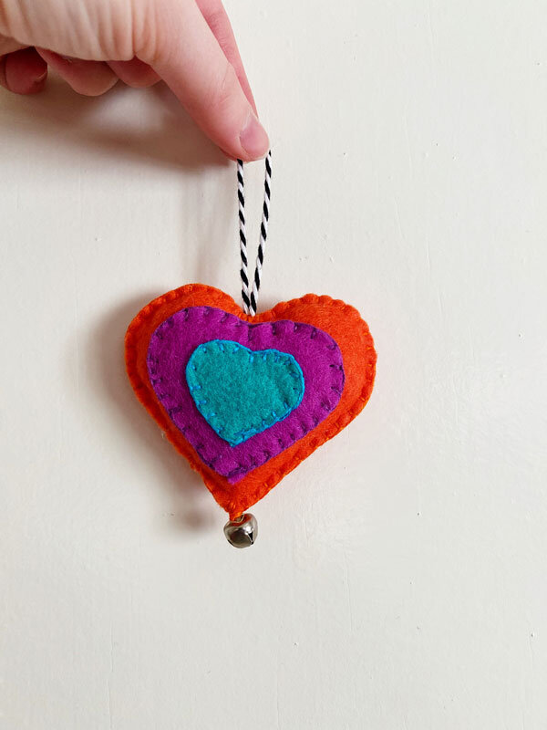 Photo of a hand holding up a colorful, hand-sewn felt heart ornament on a white background.