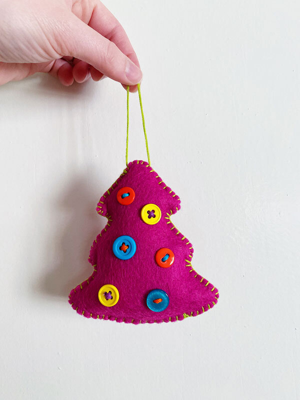 Photo of a hand holding up a fuchsia hand-sewn felt tree ornament embellished with colorful buttons on a white background.
