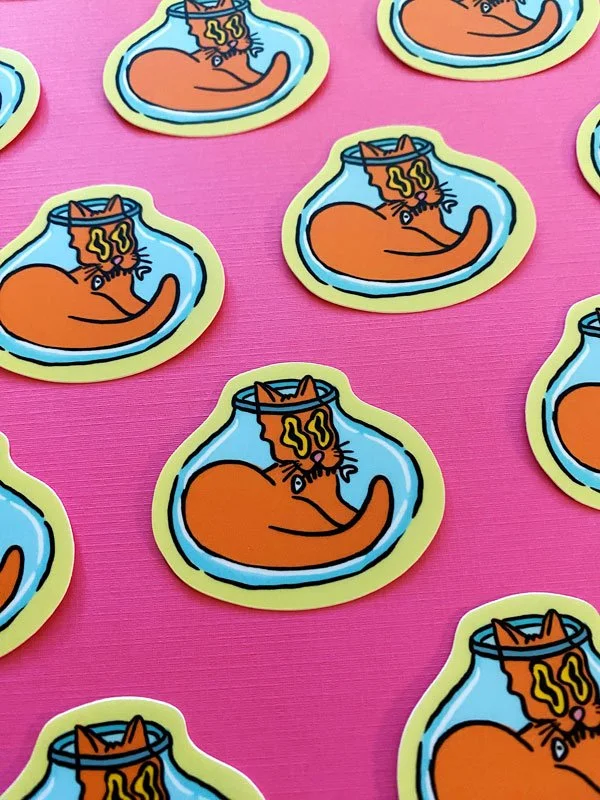 Overview group photo of vinyl stickers that depict a silly cat sitting in a fish bowl set against a pink background.