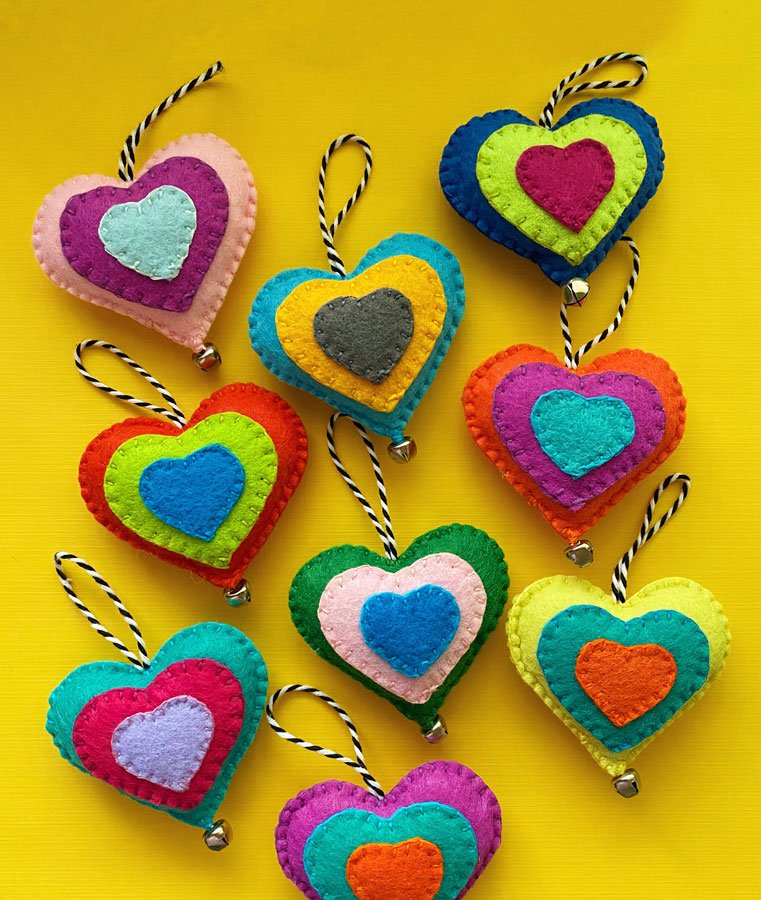 Photo of colorful, hand-sewn felt heart ornaments on a yellow background. Each heart has a jingle bell and a hanging loop.
