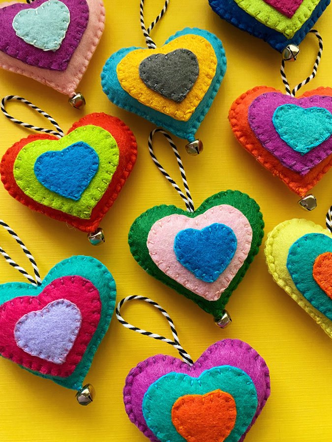 Close up of colorful, hand-sewn felt heart ornaments on a yellow background. Each heart has a jingle bell and a hanging loop.