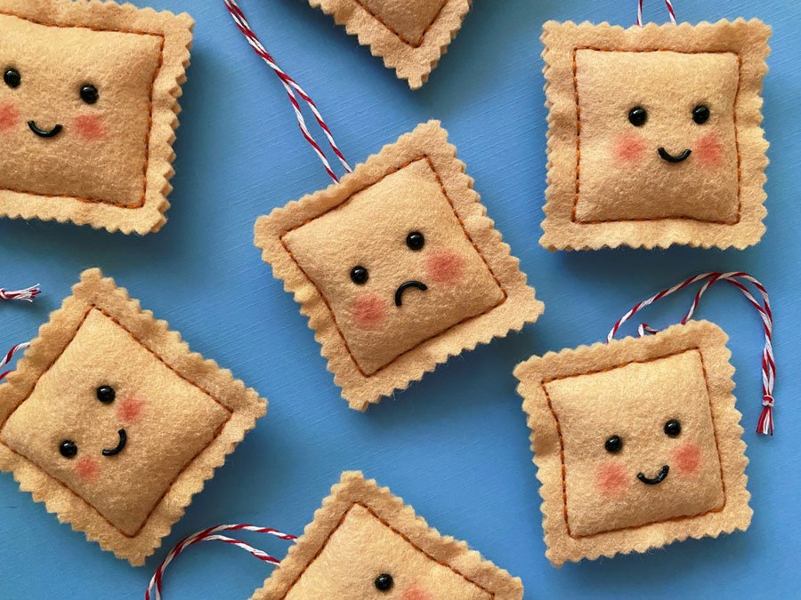 Overview photo of hand sewn felt ravioli ornaments on a blue background. Each has a happy or sad face with blushing cheeks.