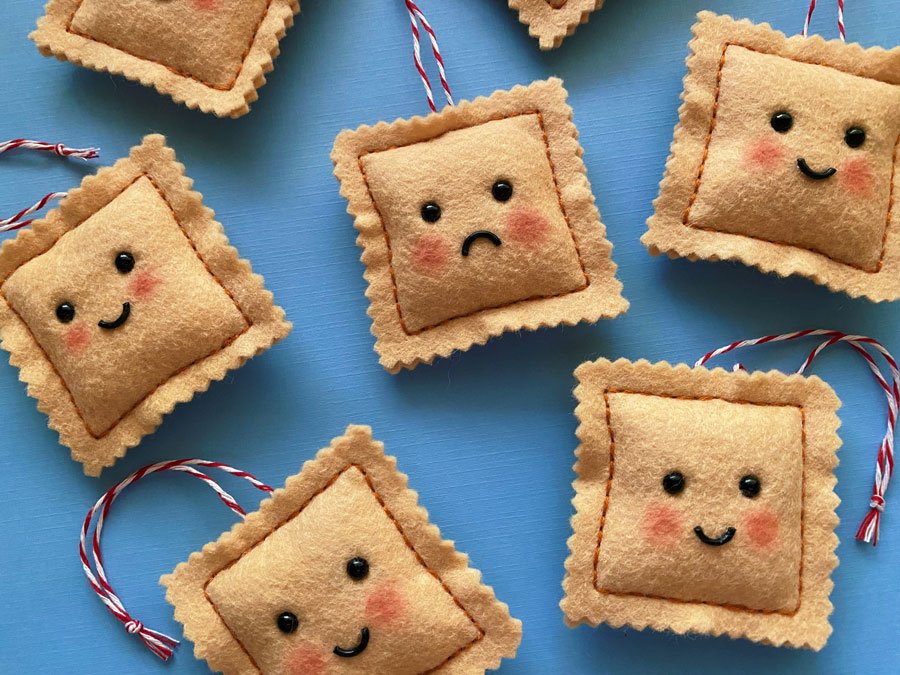 Overview photo of hand sewn felt ravioli ornaments on a blue background. Each has a happy or sad face with blushing cheeks.
