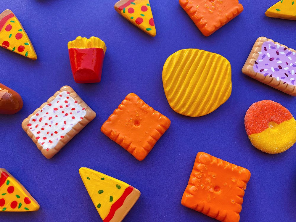 Group photo of various food magnets (such as chips, candy, pizza, and more) made out of air dry clay on blue background.