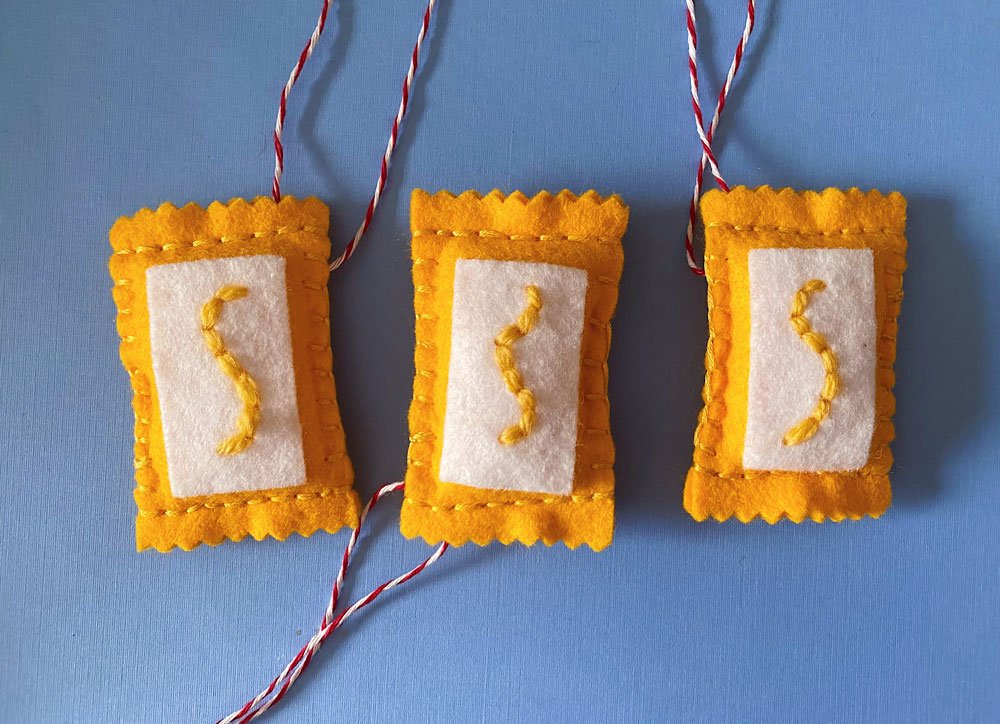 Photo of three sewn mustard ornaments on a blue background. Each mustard packet uses yellow and white felt with yellow wool.