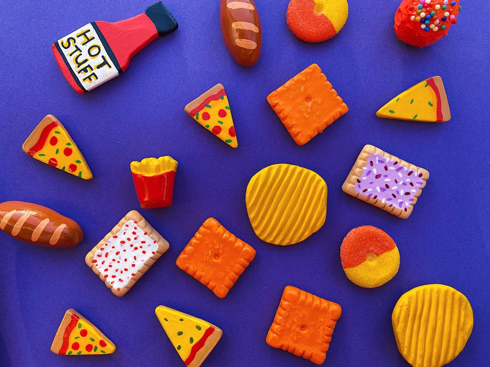 Group photo of various food magnets (such as chips, candy, pizza, and more) made out of air dry clay on blue background.