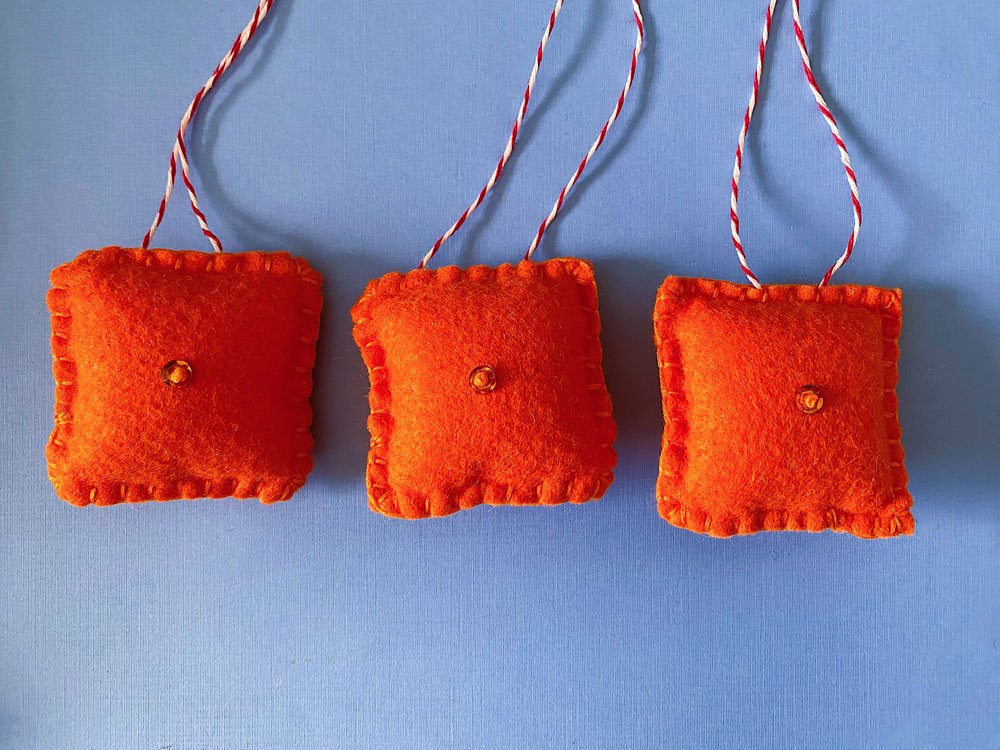 Photo of three sewn felt ornaments on a blue background. Each ornament is made with orange felt with a sequin center.