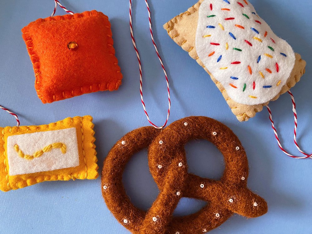 Photo of four sewn and felted food-related ornaments. Pictured is a pretzel, pop tart, cheesy cracker, and a mustard packet.