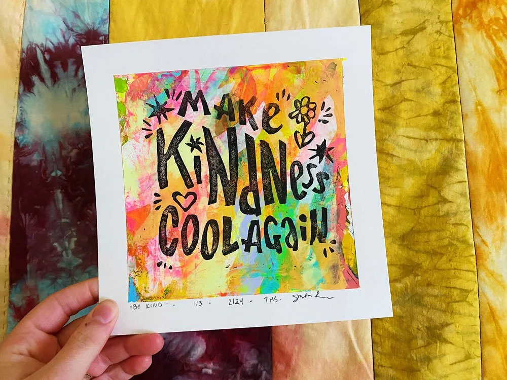Photo of a hand holding up a lino cut print with a hand-painted backing and text that reads "make kindness cool again".