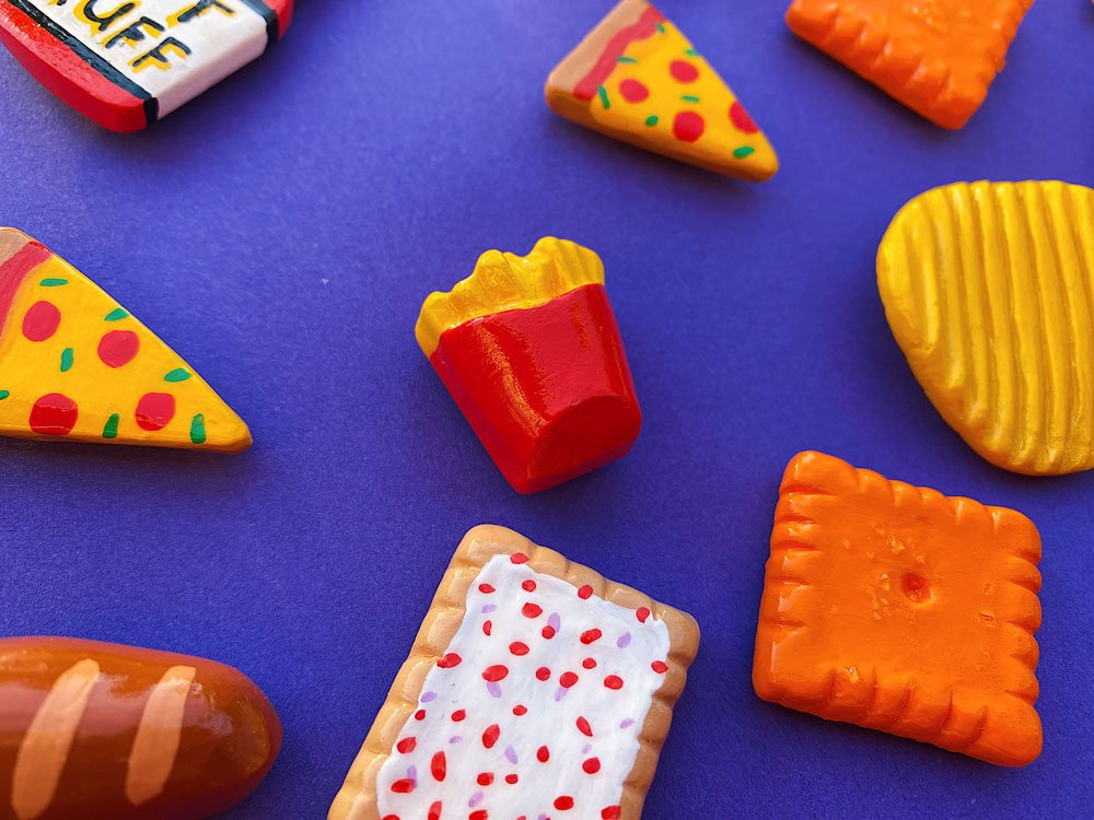 Group photo of various food magnets (such as chips, fries, pizza, bread, & more) made out of air dry clay on blue background.