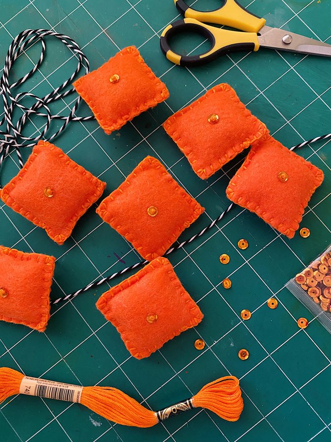 Process photo of the banner: squares of orange felt on a green cutting mat background with twine, sequins, and scissors.
