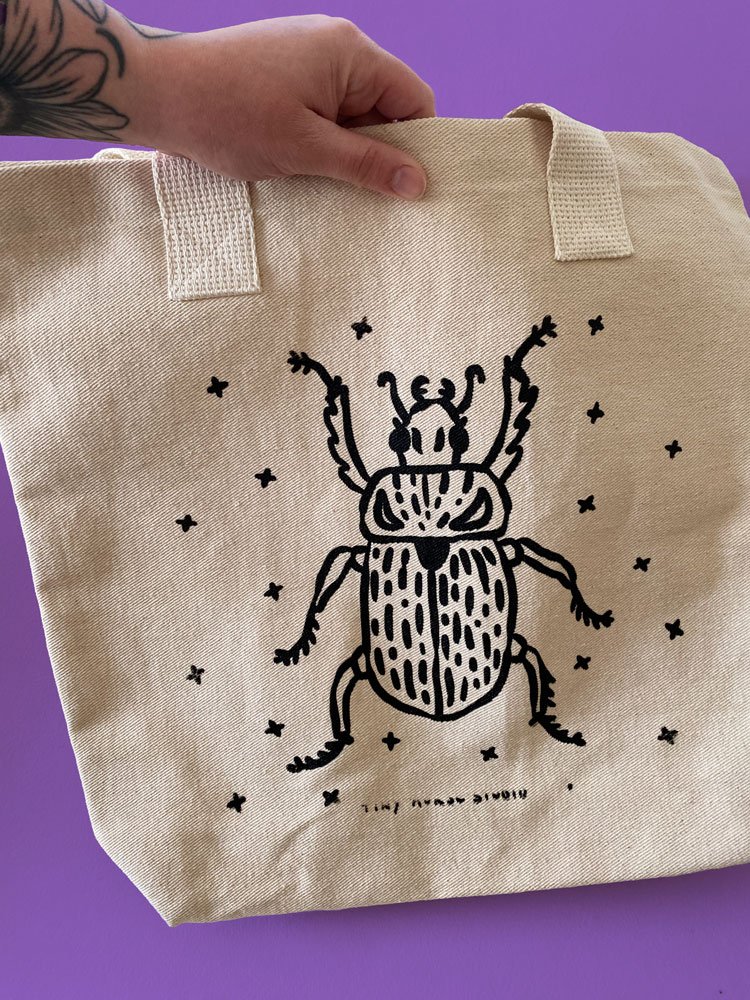 Photo of a hand holding a natural canvas tote bag with a screen printed image of a beetle on a on a purple background.