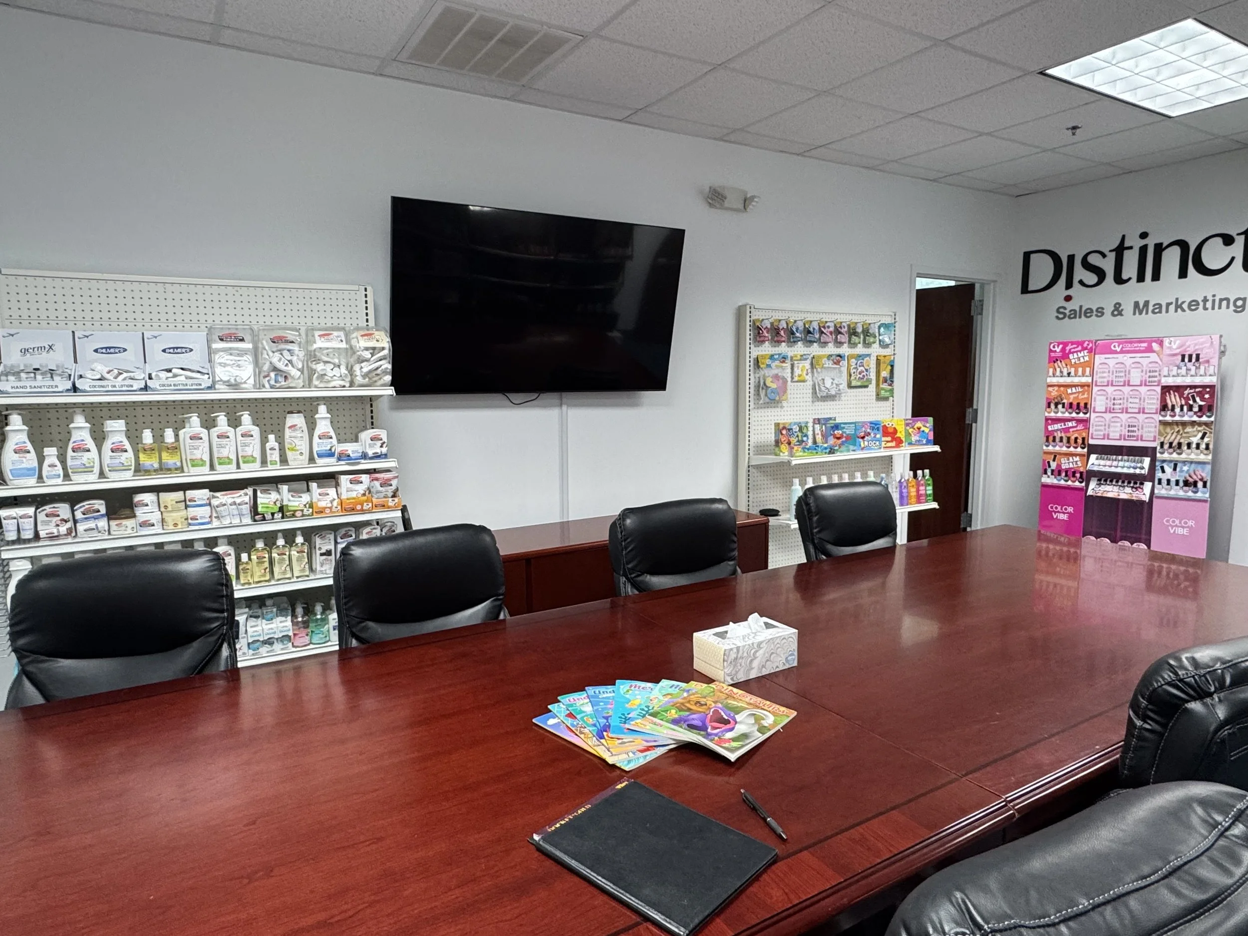 Empty conference room with black leather chairs around a large wooden table, magazines and a pen on the table, shelves with products, a large TV screen on the wall, and a pink display stand with nail polish bottles.