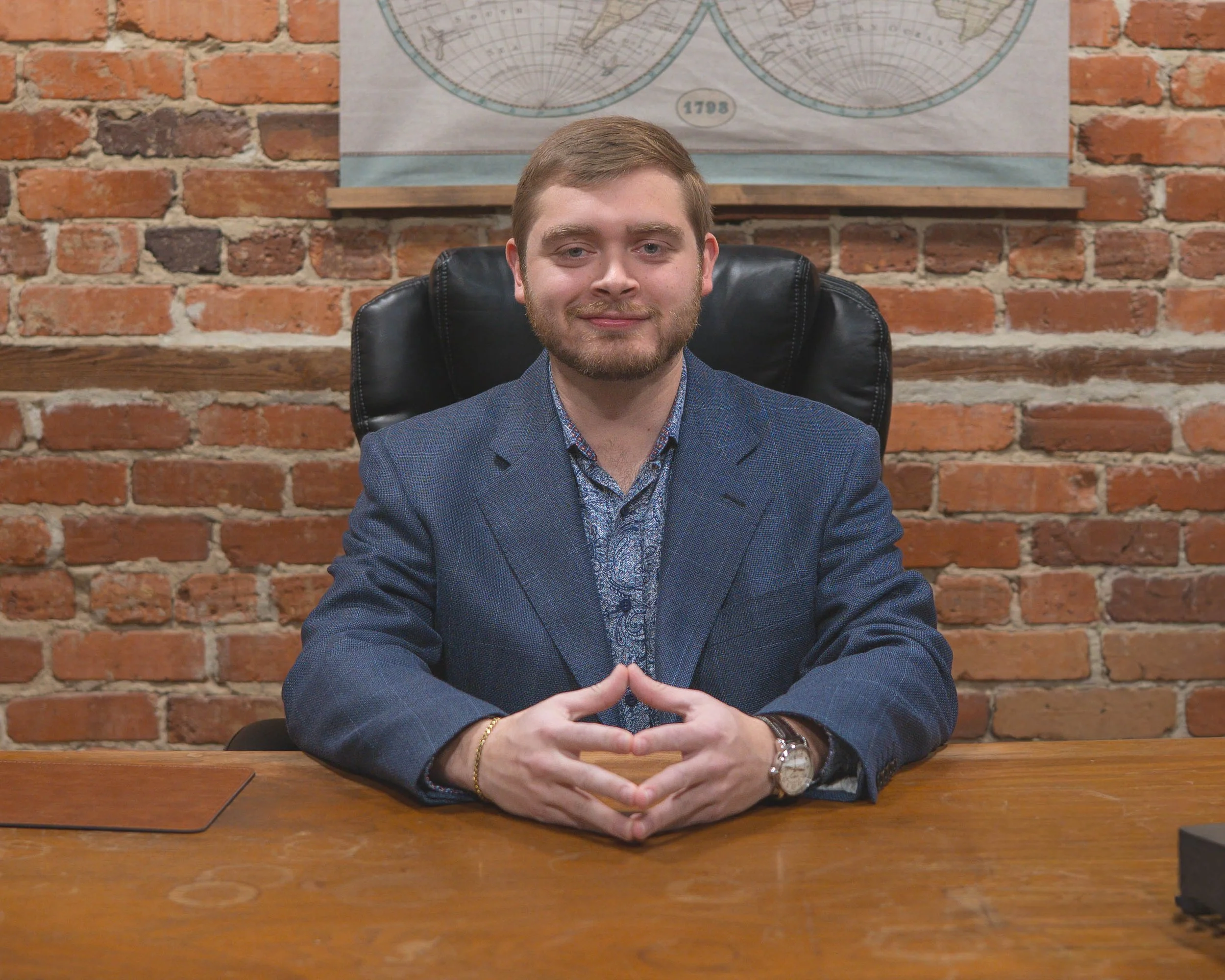A real estate broker with light brown hair and beard, wearing a dark blue blazer and patterned shirt, sitting at a wooden table with a brick wall background and a world map poster behind him.