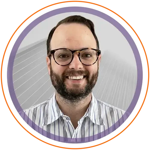 A man with glasses and a beard, smiling, wearing a striped shirt, in front of a gray curved wall.