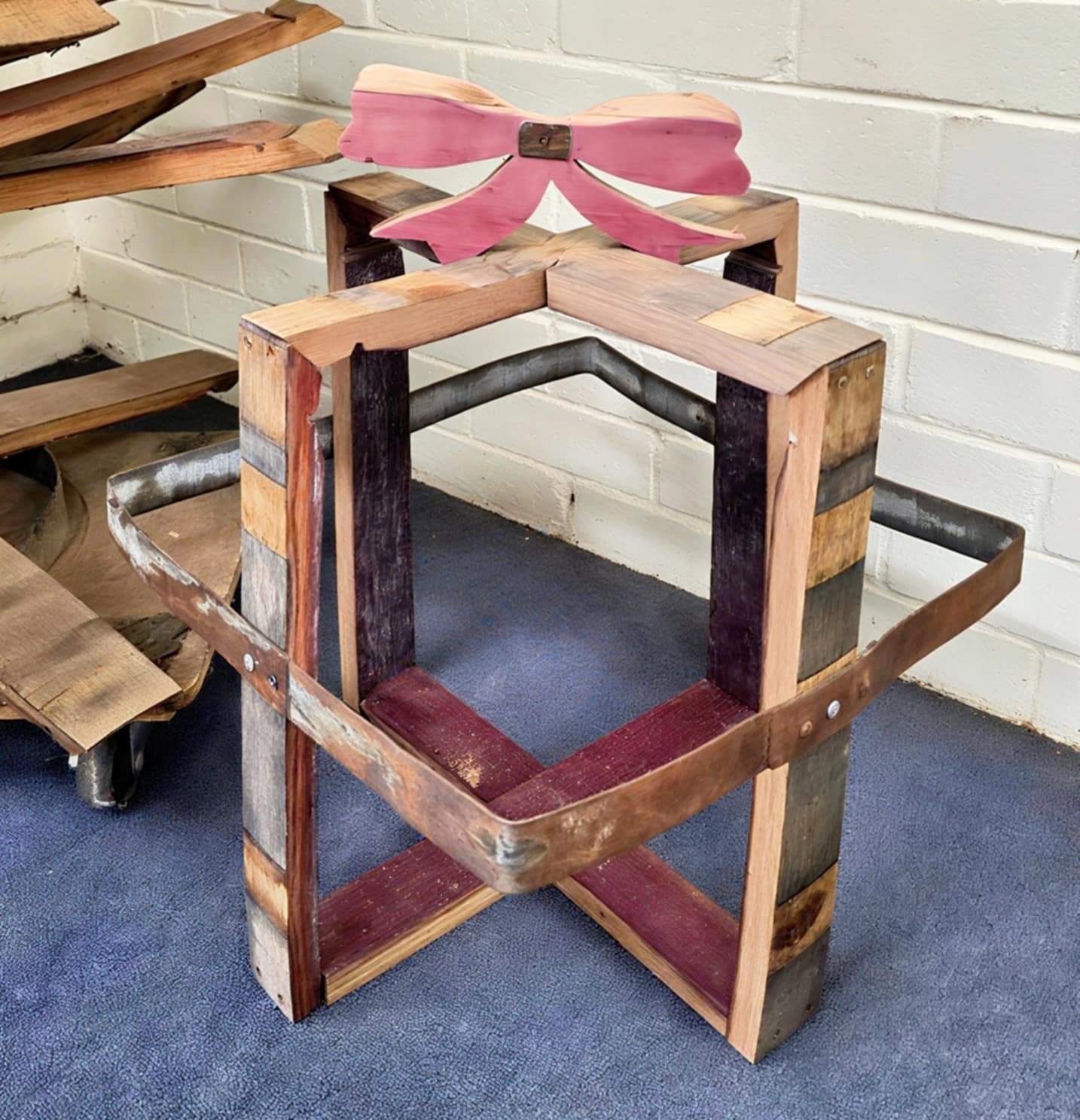 A wooden chair frame with a pink wooden bow on top, placed on blue carpet near a white brick wall.