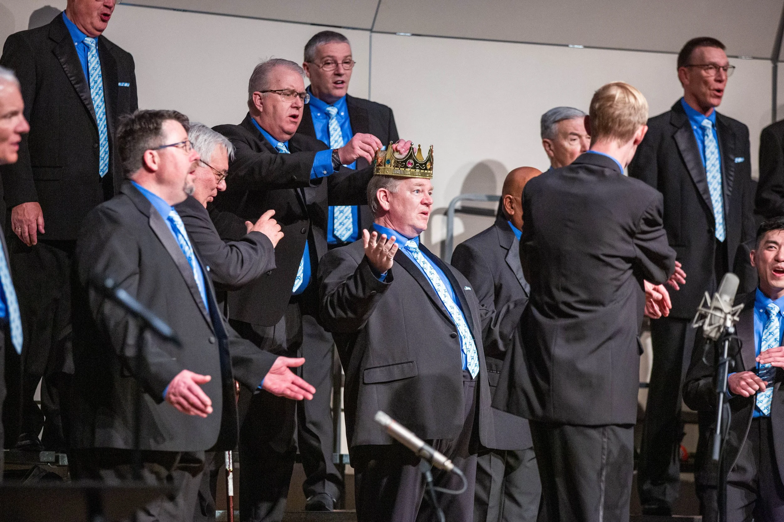 Members of Acappella Road performing onstage in black suits with blue shirts and patterned ties. One member in the center of the frame is adorning another member's head with a gold plastic crown.