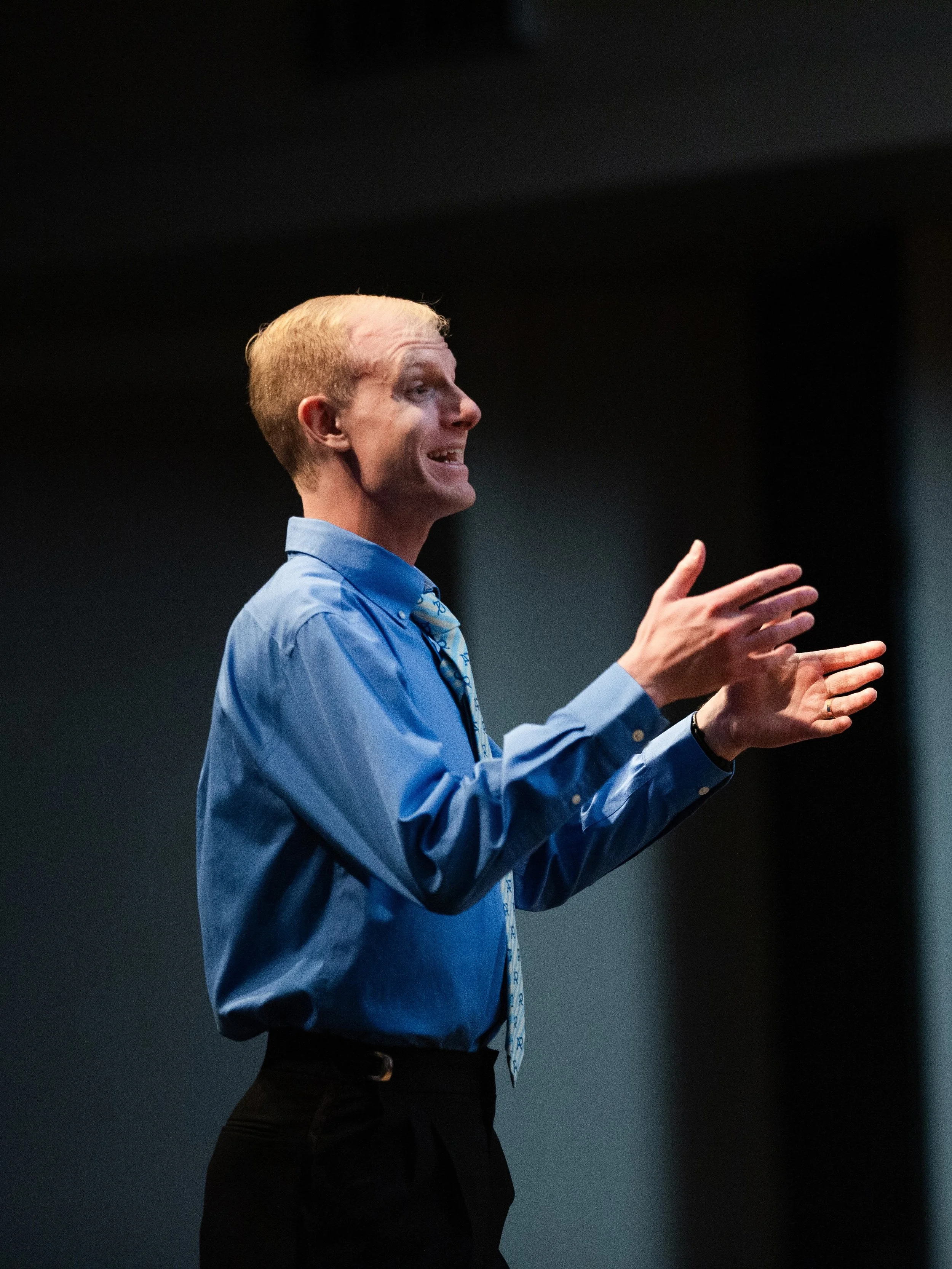 Acappella Road's director Adrian Smith, dressed in a blue shirt and patterned tie, in front of a black backdrop. He is smiling with his hands raised in the midst of conducting a song.