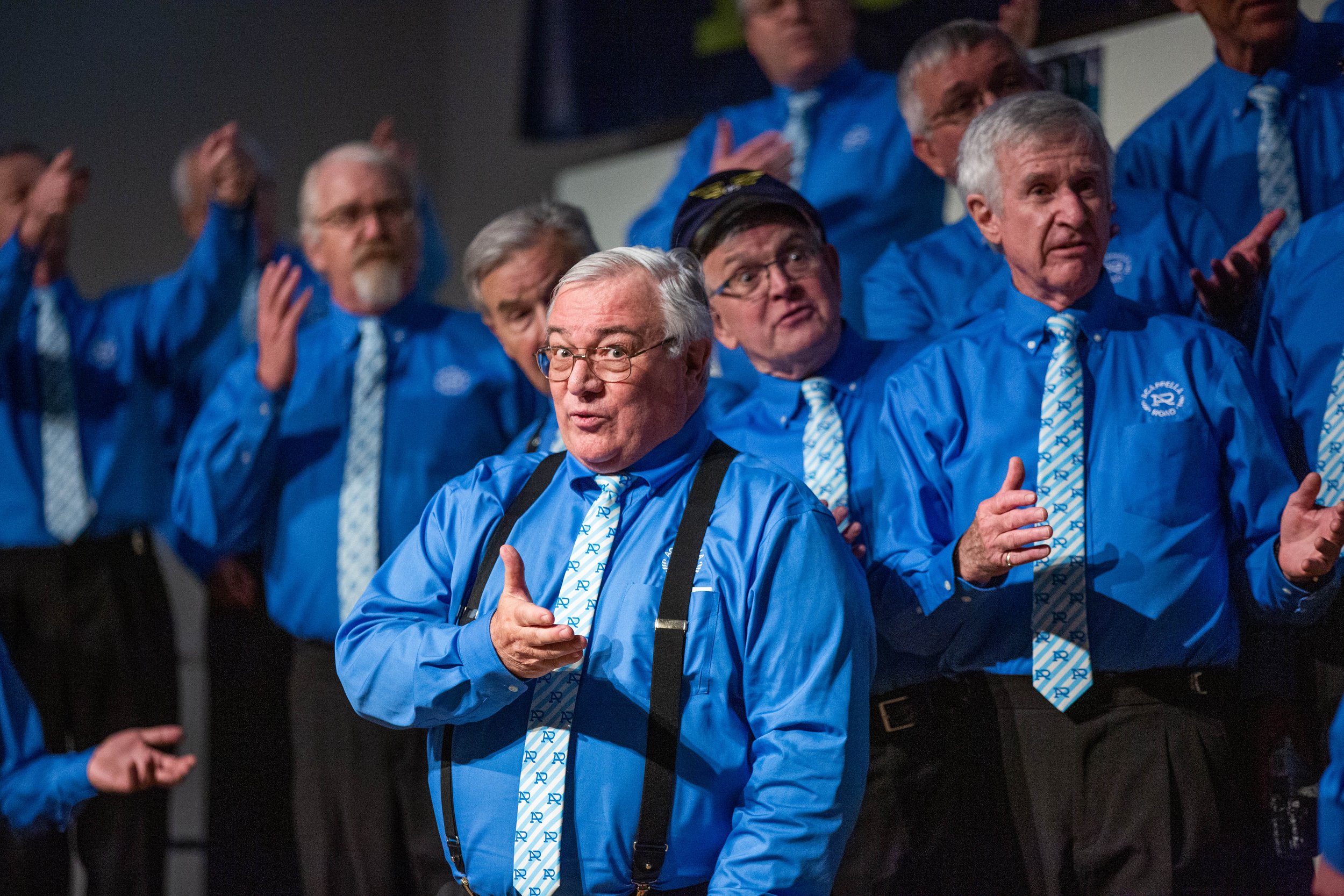 Members of Acappella Road performing onstage. One member in the foreground has a hand outstretched and is looking at the camera with an expectant expression.