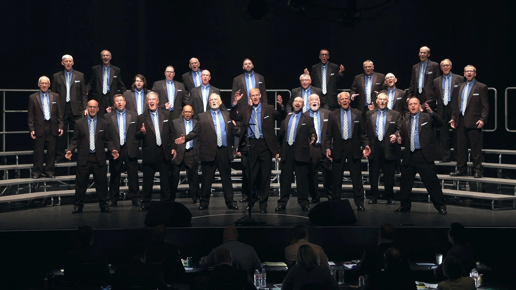 Acappella Road chorus pictured performing onstage in black suits with blue shirts and ties, posing at the end of a song.