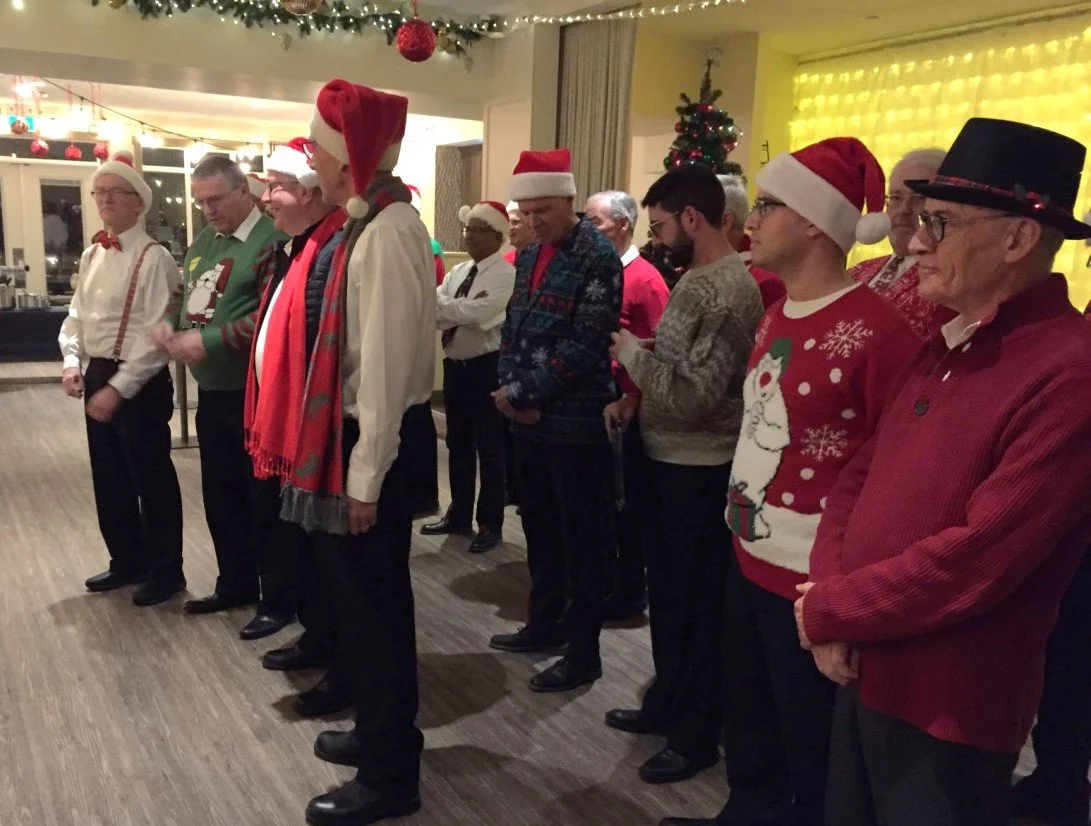 About a dozen members of Acappella Road standing ready to perform, wearing various Christmas-themed attire.