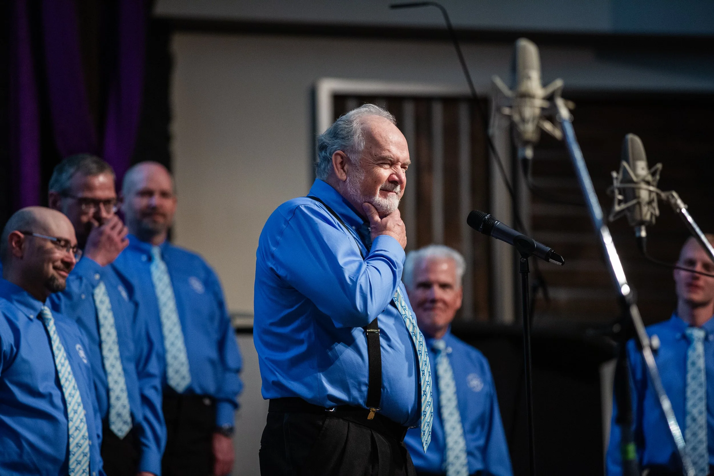 Members of Acappella Road pictured onstage wearing blue shirts and patterned ties. The member in the foreground is standing at a microphone and smiling, holding his chin with a cheeky expression.