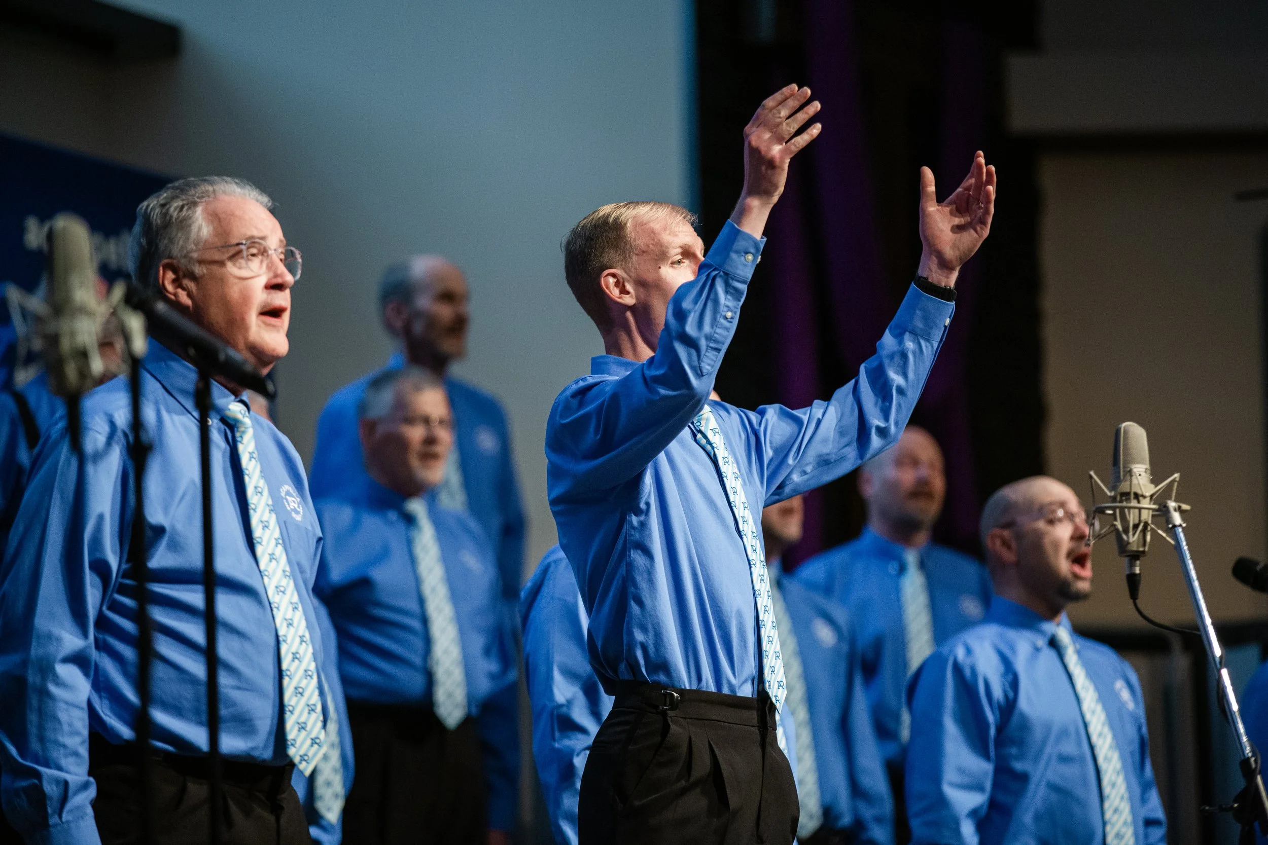 Members of Acappella Road performing onstage in blue uniforms with patterned ties. The director, Adrian Smith, is in the foreground with his arms raised.
