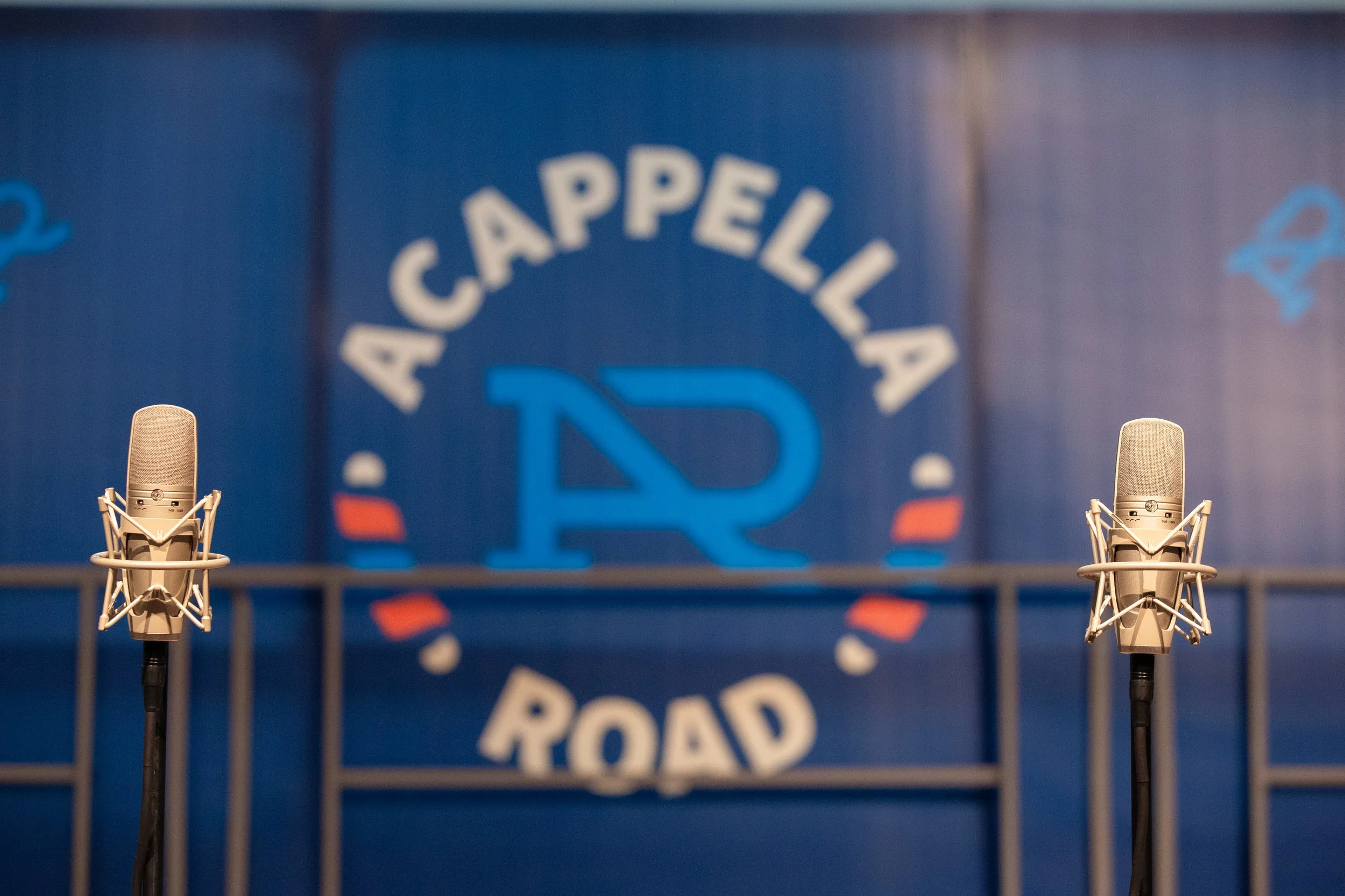 Acappella Road's logo pictured on a concert backdrop, with two microphones in the foreground to the left and right sides.