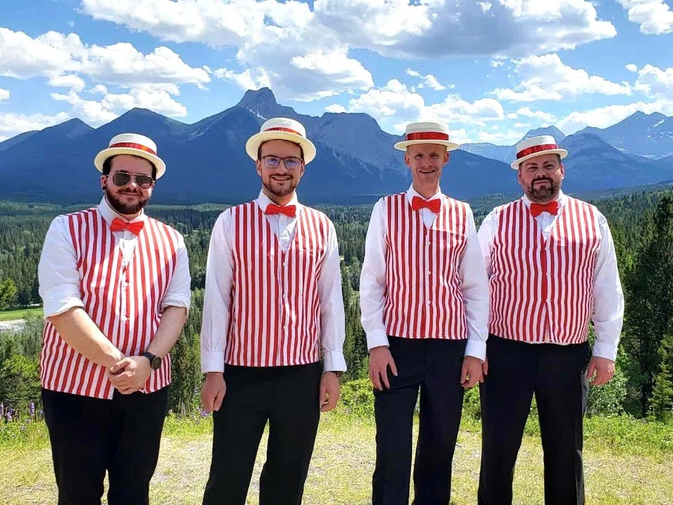 Major Inconvienience, a four-man barbershop quartet, posing outdoors in front of a mountain range, wearing traditional striped vests and boater hats.