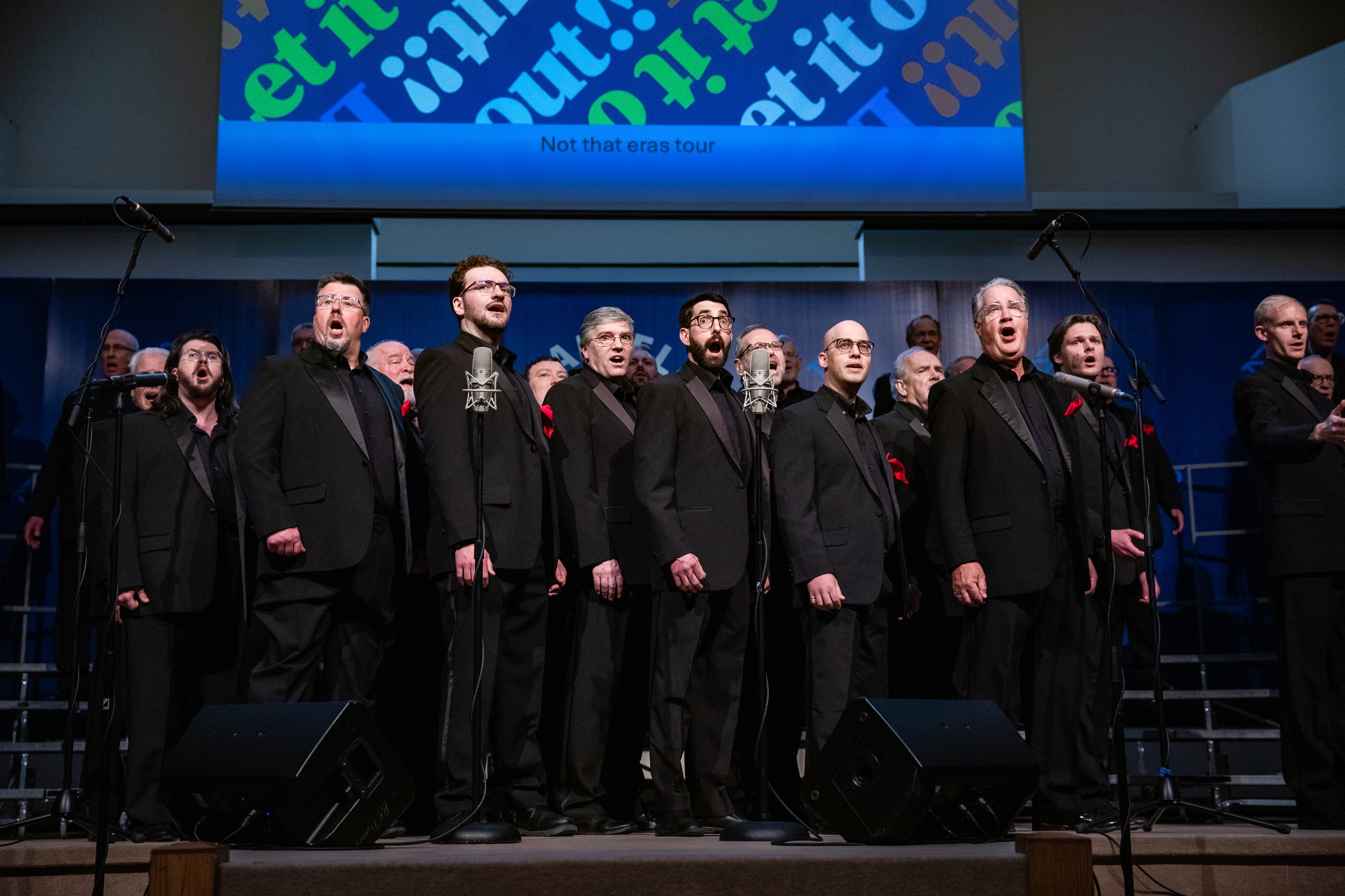 Acappella Road members performing in concert in black suits with red pocket squares, singing a chord together with mouths open wide.