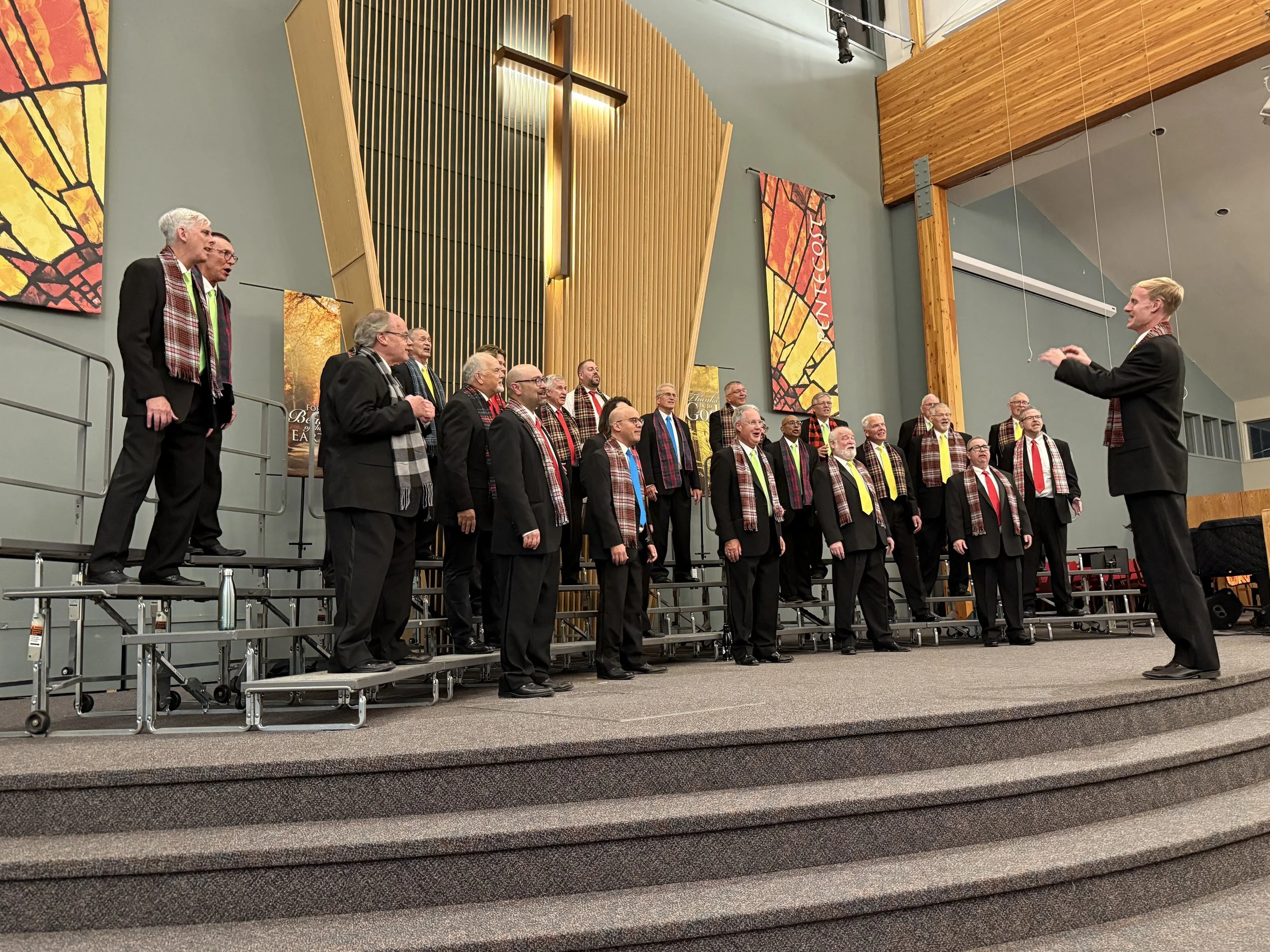 Members of Acappella Road performing onstage at McDougall United Church. They are wearing black suits, brightly coloured ties, and plaid scarves in a variety of brown tones.